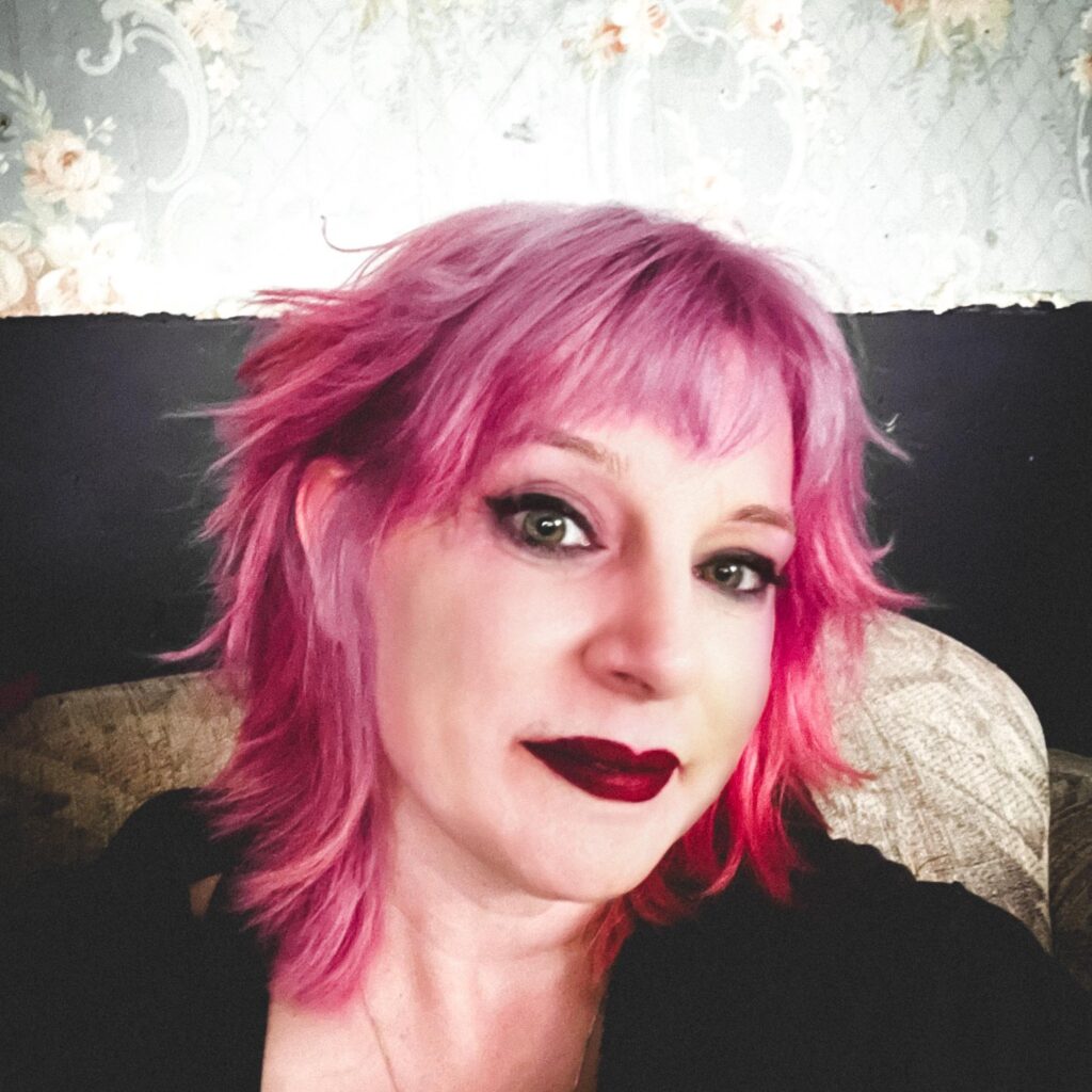 Portrait of a woman with vibrant pink hair and deep berry lipstick, softly smiling. She has a grounded yet radiant presence, captured in a cozy, vintage-inspired setting.